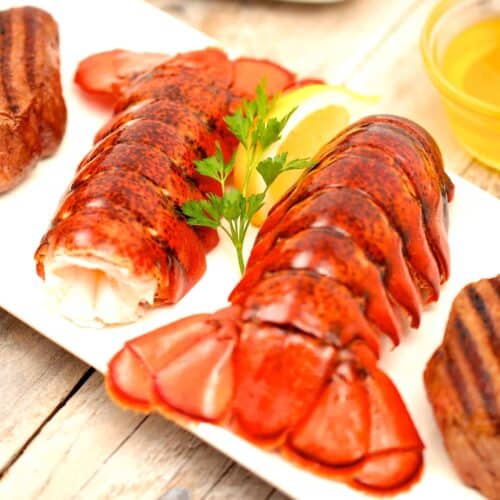 Succulent grilled lobster tails served on a white plate alongside perfectly seared steak, fresh parsley, and lemon garnish. A premium seafood dish featuring tender, buttery lobster meat, ideal for gourmet dining. Perfect for those looking to buy lobster tails online for a luxurious seafood experience at home.