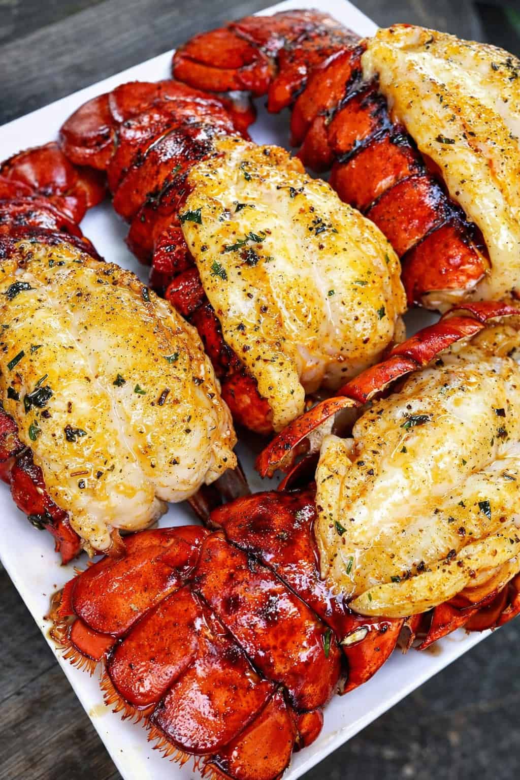 Grilled lobster tails topped with lemon butter sauce and herbs on a serving platter.