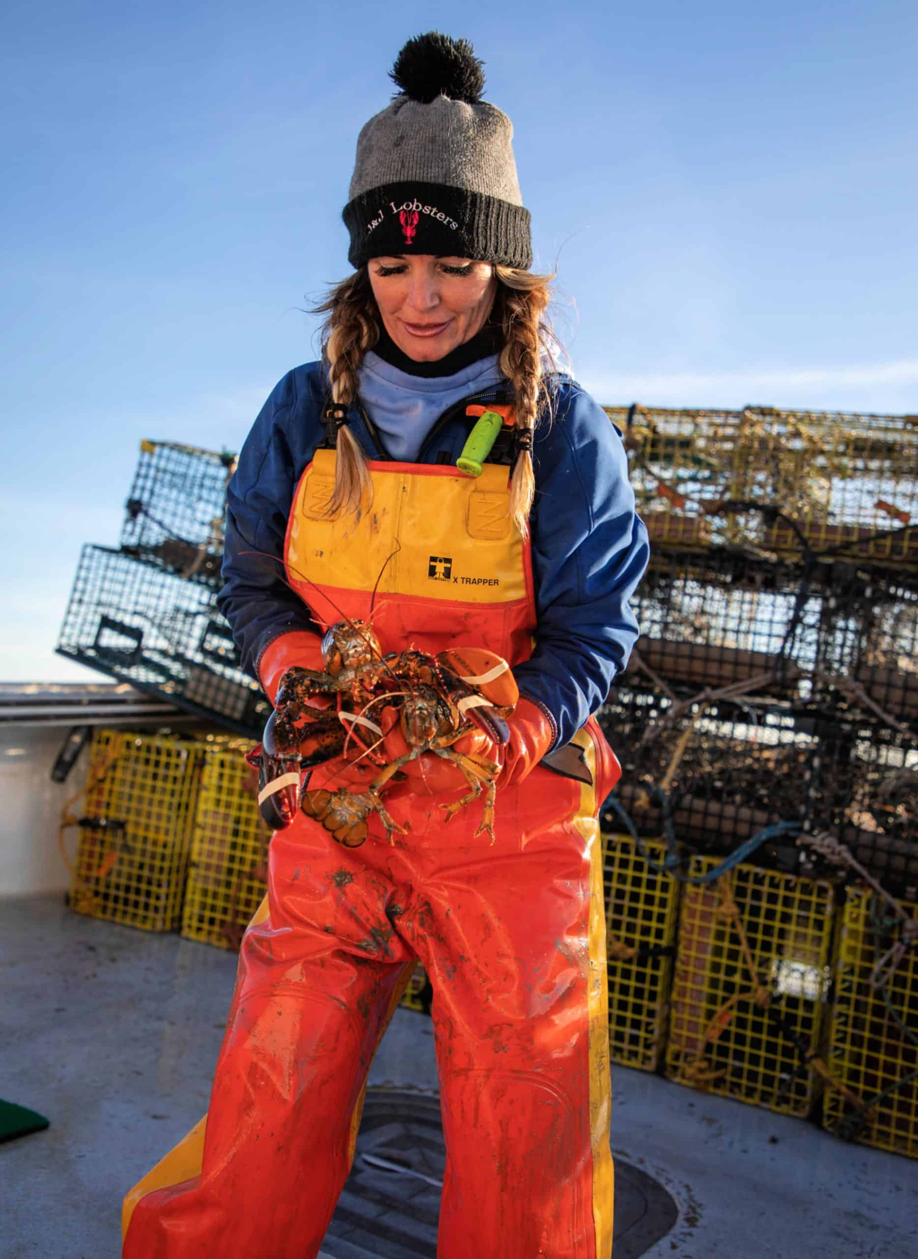 Maine lobsterwoman in orange oilskins holding two hard-shell lobsters on a boat stacked with traps