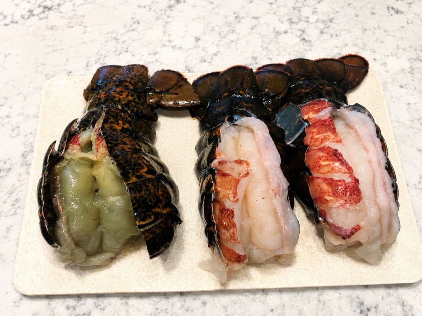 Three split raw lobster tails on a cutting board, one with green-tinted meat and two with normal white meat.