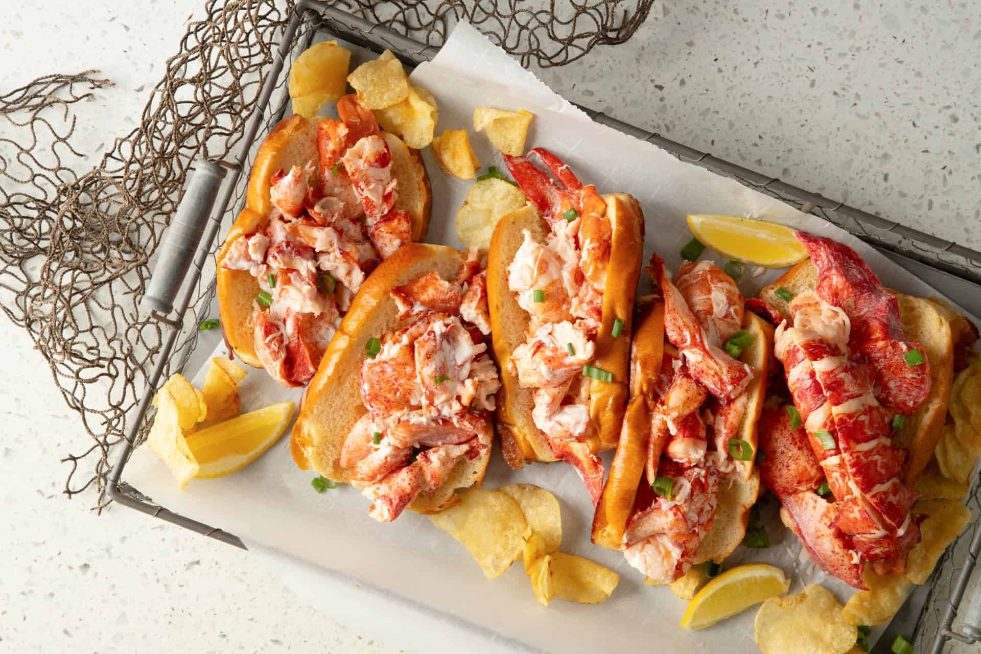 Lobster rolls on toasted top-split buns overflowing with claw, knuckle, and tail meat, garnished with chives, served with lemon wedges and potato chips