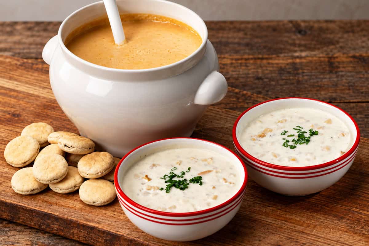 Lobster bisque in a large white bowl with two smaller bowls of clam chowder and oyster crackers on a rustic wooden table