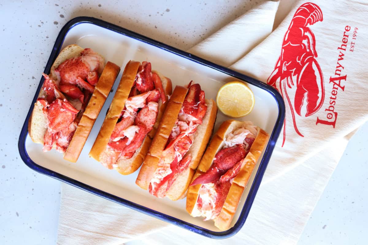 Four lobster rolls on a white serving tray with lemon and a LobsterAnywhere towel