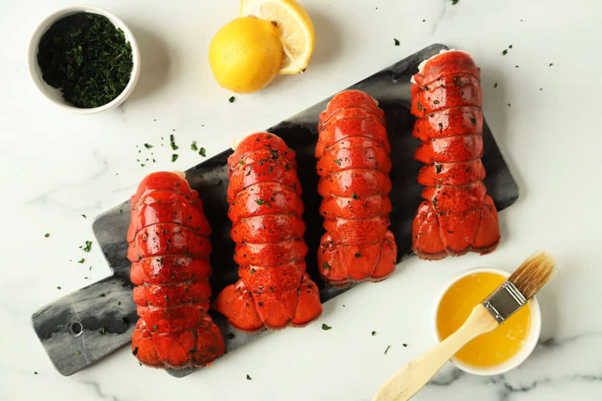 Four cooked lobster tails on a serving board with lemon, herbs, and melted butter