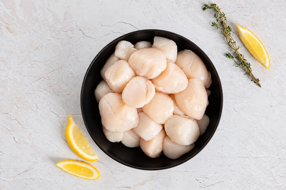 Raw sea scallops in a black bowl with lemon wedges and thyme on a light stone surface